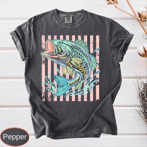 Puede incluir: Camiseta gris oscuro con una ilustraci&oacute;n colorida de un pez con la boca abierta, sobre un fondo de rayas rosas. El pez tiene detalles azules, amarillos y verdes. La camiseta tiene las mangas enrolladas.