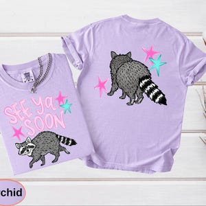 May include: Two orchid-colored t-shirts. One shirt features the text "SEE YA SOON" with a raccoon graphic and pink and teal stars. The other shirt has a raccoon graphic and pink and teal stars on the back.