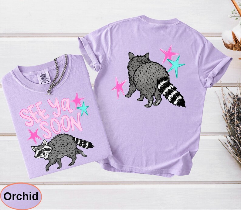 May include: Two orchid-colored t-shirts. One shirt features the text "SEE YA SOON" with a raccoon graphic and pink and teal stars. The other shirt has a raccoon graphic and pink and teal stars on the back.