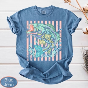 Puede incluir: Camiseta azul vaquera con una ilustraci&oacute;n colorida de un pez sobre un fondo de rayas rosas. El pez est&aacute; representado en tonos verdes, azules, amarillos y morados. La camiseta tiene cuello redondo y mangas cortas. La tela es suave.