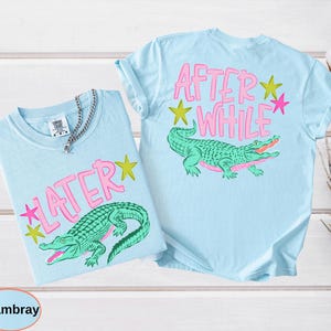Puede incluir: Dos camisetas azul claro con las palabras "LATER" y "AFTER WHILE" en rosa, junto con un gr&aacute;fico de cocodrilo verde. Las camisetas tambi&eacute;n presentan acentos de estrellas rosas y amarillas. Una camiseta tiene un collar de cadena plateada.