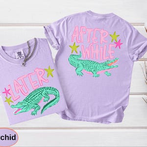 Puede incluir: Dos camisetas color orqu&iacute;dea con dise&ntilde;os a juego. Cada camiseta presenta un gr&aacute;fico de cocodrilo verde con el texto "LATER" y "AFTER WHILE" en rosa. Estrellas amarillas y rosas acent&uacute;an el dise&ntilde;o.