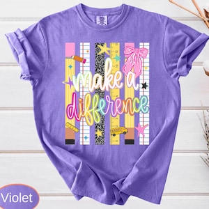 May include: A violet t-shirt featuring a graphic print with the text "make a difference" in colorful script. The design includes vertical stripes, a yellow ruler, a pink bow, a small yellow school bus, and various star shapes in a school theme.