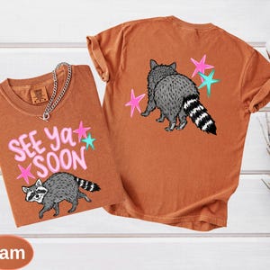 May include: Two orange-brown t-shirts with raccoon graphics and the text "See Ya Soon." One shirt shows the raccoon from the front, the other from the back. Pink and teal star accents are included in the design. A silver chain necklace is draped on the front shirt.