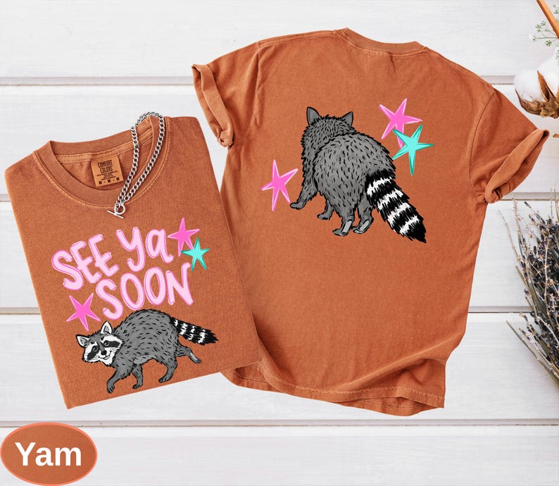 May include: Two orange-brown t-shirts with raccoon graphics and the text "See Ya Soon." One shirt shows the raccoon from the front, the other from the back. Pink and teal star accents are included in the design. A silver chain necklace is draped on the front shirt.