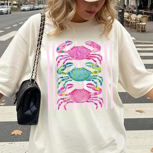 May include: Cream-colored t-shirt featuring a colorful crab design. The graphic includes three crabs in pink, blue, and green, with pink vertical stripes. The shirt is a casual, everyday wear item.