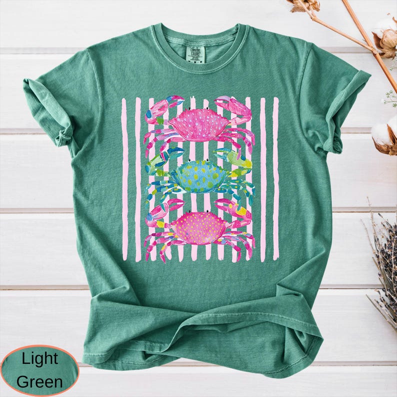 May include: Light green t-shirt featuring a colorful crab design. The design includes three crabs in pink and blue, set against a backdrop of vertical pink stripes. The shirt is made of a soft material.