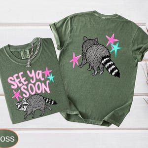 May include: Two moss green t-shirts with raccoon graphics and the text "See Ya Soon". One shirt shows the raccoon walking, the other shows the raccoon from behind. Pink and teal stars accent the design. A silver chain necklace is on one shirt.