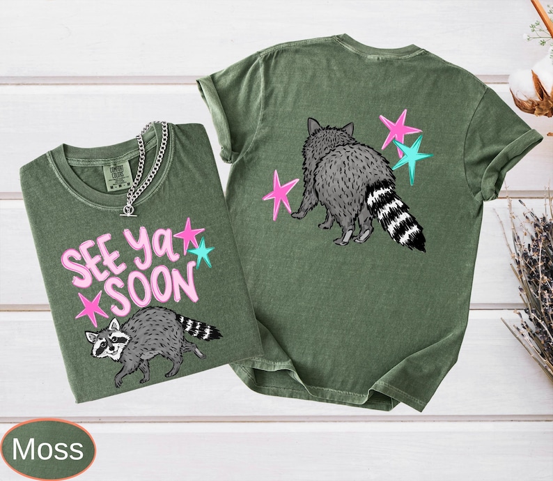 May include: Two moss green t-shirts with raccoon graphics and the text "See Ya Soon". One shirt shows the raccoon walking, the other shows the raccoon from behind. Pink and teal stars accent the design. A silver chain necklace is on one shirt.