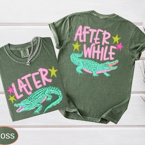 Puede incluir: Dos camisetas verde musgo con gr&aacute;ficos de caimanes. Una camiseta tiene "LATER" y la otra "AFTER WHILE" en texto rosa con acentos de estrellas amarillas y rosas. Una cadena de plata est&aacute; en la primera camiseta.