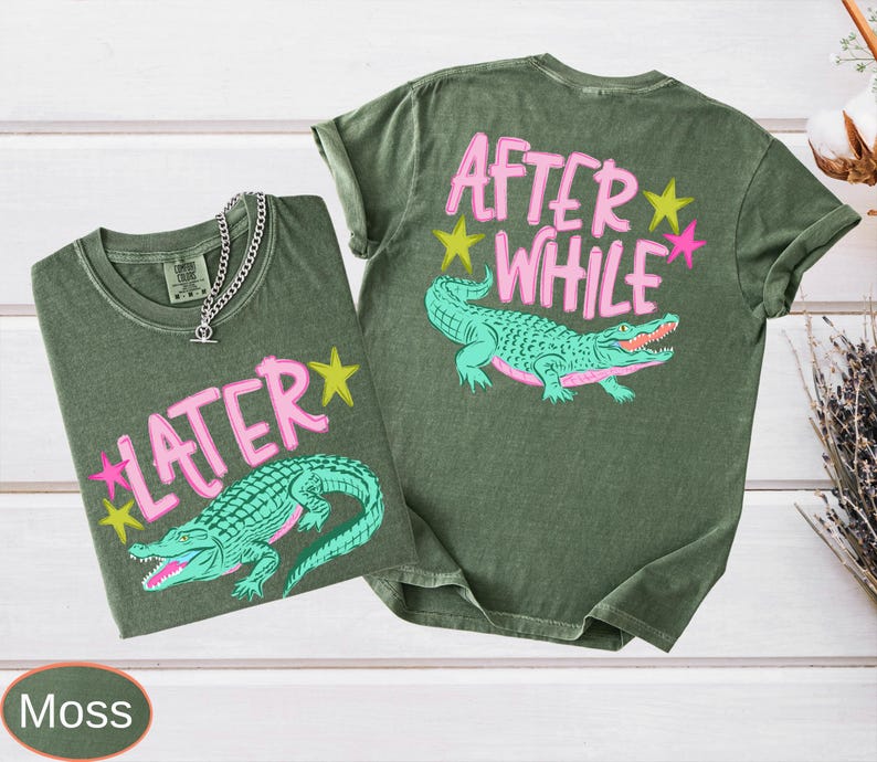 Puede incluir: Dos camisetas verde musgo con gr&aacute;ficos de caimanes. Una camiseta tiene "LATER" y la otra "AFTER WHILE" en texto rosa con acentos de estrellas amarillas y rosas. Una cadena de plata est&aacute; en la primera camiseta.