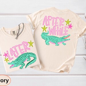 Puede incluir: Dos camisetas color marfil con dise&ntilde;os de caimanes. Una camiseta dice "LATER" con una estrella rosa, y la otra dice "AFTER WHILE" con una estrella rosa. Los caimanes son turquesas con detalles rosas.