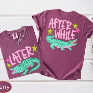 Puede incluir: Dos camisetas color bayas con dise&ntilde;os gr&aacute;ficos. Una camiseta presenta el texto "LATER" con un caim&aacute;n verde y estrellas. La otra camiseta tiene "AFTER WHILE" con un caim&aacute;n y estrellas.