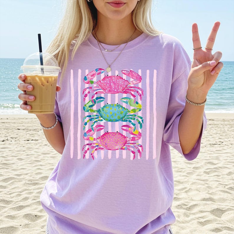 May include: A light purple t-shirt featuring a vertical striped design with three colorful crab illustrations. The crabs are pink, blue, and green with detailed watercolor patterns. The shirt is a casual, oversized fit.