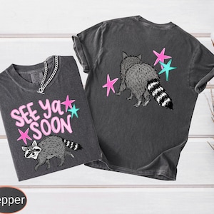 May include: Two dark gray t-shirts with raccoon graphics and the text "See Ya Soon." One shirt has the text and raccoon on the front, the other has a raccoon and stars on the back. The raccoon is gray and white with a striped tail.