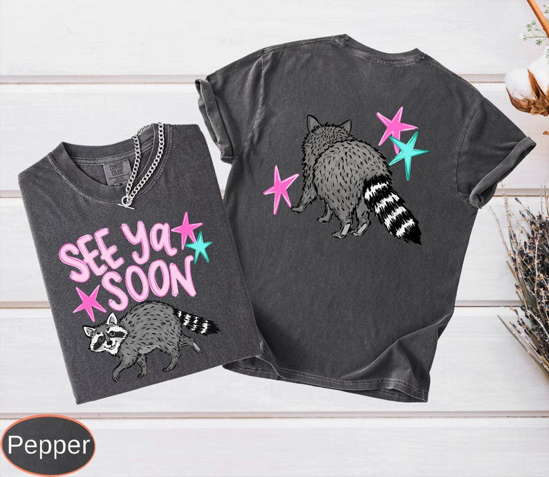 May include: Two dark gray t-shirts with raccoon graphics and the text "See Ya Soon." One shirt has the text and raccoon on the front, the other has a raccoon and stars on the back. The raccoon is gray and white with a striped tail.