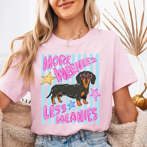 May include: Pink t-shirt with a graphic of a dachshund wearing heart-shaped glasses. The text reads "MORE WEENIES LESS MEANIES" with star accents. The shirt has a crew neck and short sleeves.