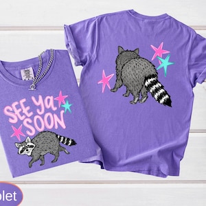 Two violet t-shirts with raccoon graphics. One shirt has the text "SEE YA SOON" with pink and teal stars. The other shirt features a raccoon and pink and teal stars on the back.