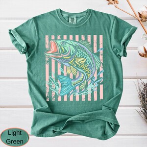 Puede incluir: Camiseta verde claro con un dise&ntilde;o de pez colorido. El pez est&aacute; representado en tonos azules, amarillos y morados, sobre un fondo de rayas rosas verticales. La camiseta tiene un corte relajado.