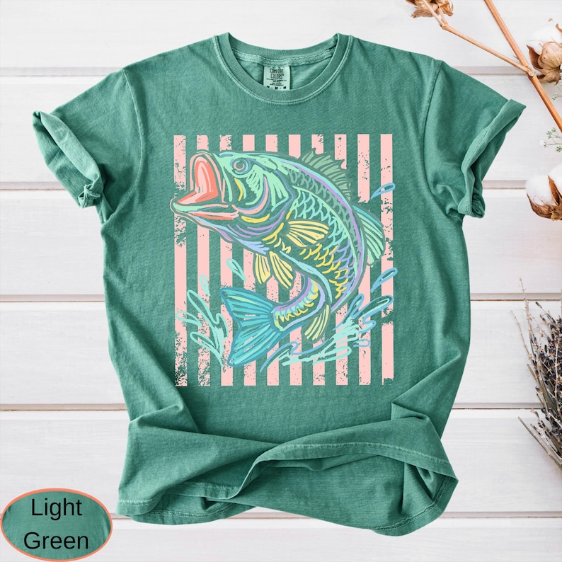 Puede incluir: Camiseta verde claro con un dise&ntilde;o de pez colorido. El pez est&aacute; representado en tonos azules, amarillos y morados, sobre un fondo de rayas rosas verticales. La camiseta tiene un corte relajado.