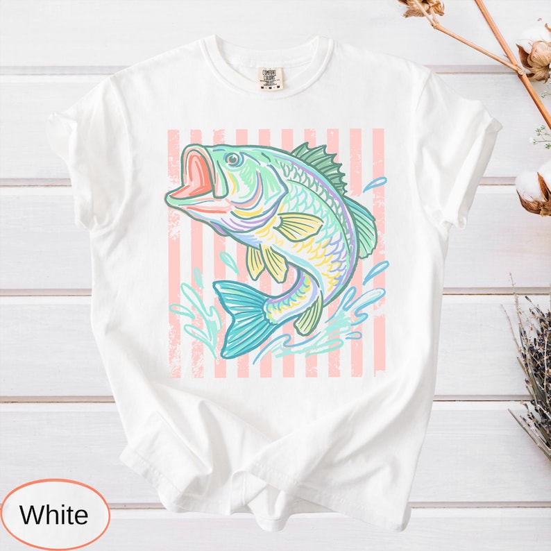 Puede incluir: Camiseta blanca con una ilustraci&oacute;n colorida de un pez saltando. El pez est&aacute; representado en tonos de azul, verde, amarillo y rosa, sobre un fondo a rayas rosas y blancas. La camiseta es de estilo casual.