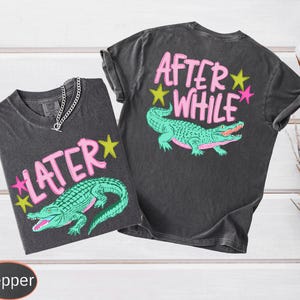 Puede incluir: Dos camisetas gris carb&oacute;n con un gr&aacute;fico de un caim&aacute;n verde y los textos "LATER" y "AFTER WHILE" en rosa. Las camisetas tienen cuello redondo y mangas cortas. Una cadena de plata est&aacute; en una camiseta.
