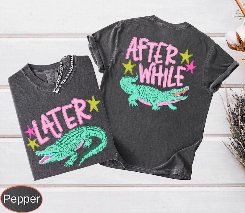 Puede incluir: Dos camisetas gris carb&oacute;n con un gr&aacute;fico de un caim&aacute;n verde y los textos "LATER" y "AFTER WHILE" en rosa. Las camisetas tienen cuello redondo y mangas cortas. Una cadena de plata est&aacute; en una camiseta.