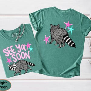May include: Two light green t-shirts with raccoon graphics. One shirt has the text "SEE YA SOON" with pink and teal stars. The other shirt features a raccoon with pink and teal stars. The shirts are made of a soft material.