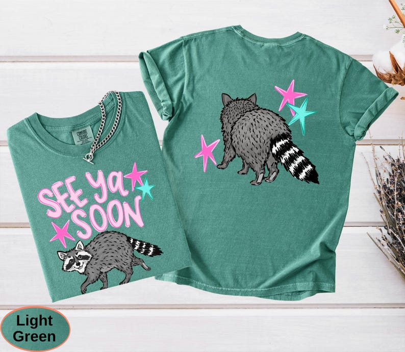 May include: Two light green t-shirts with raccoon graphics. One shirt has the text "SEE YA SOON" with pink and teal stars. The other shirt features a raccoon with pink and teal stars. The shirts are made of a soft material.