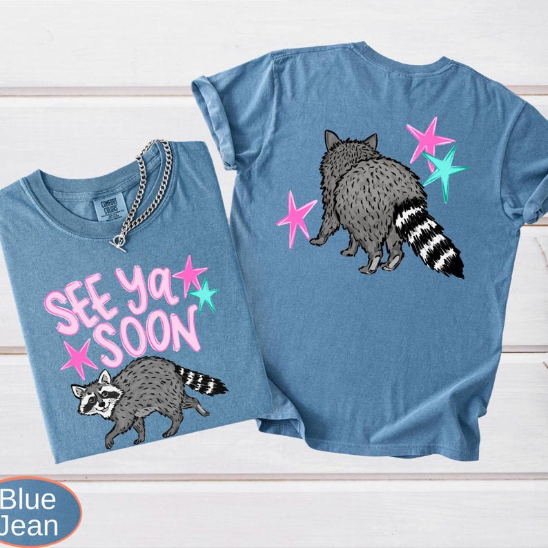 Product photography displaying the front and back of a dark grey shirt with a raccoon design. The front has the 'SEE YA SOON' pink text above a walking raccoon and colorful stars. The back has the rear view of the raccoon walking away with the striped tail visible, the same text, and more stars.