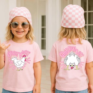 May include: Pink t-shirt with the text "Guess What?" and a cartoon chicken on the front. The back of the shirt features the text "Chicken Butt" and a cartoon chicken. The child is wearing a pink and white checkered beanie.