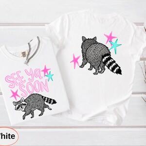 May include: Two white t-shirts with raccoon graphics. One shirt has the text "SEE YA SOON" in pink with star accents. The other shirt features a raccoon with star accents. Both shirts have a crew neck.