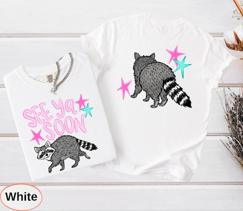 May include: Two white t-shirts with raccoon graphics. One shirt has the text "SEE YA SOON" in pink with star accents. The other shirt features a raccoon with star accents. Both shirts have a crew neck.