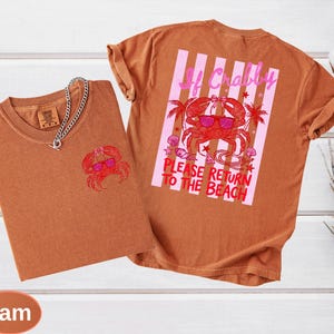 May include: Two yam-colored t-shirts with a crab graphic. One shirt has a small crab design on the front, and the other has a larger crab design with text that reads "If Crabby, Please Return to the Beach". A silver chain necklace is on the front shirt.