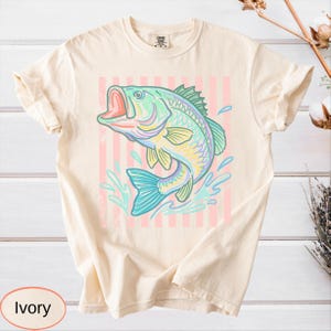 Puede incluir: Camiseta de color marfil con una ilustraci&oacute;n colorida de un pez saltando con la boca abierta, salpicaduras de agua y un fondo a rayas rosas. El pez es en tonos de azul, verde, amarillo y morado. La camiseta tiene cuello redondo y mangas cortas.