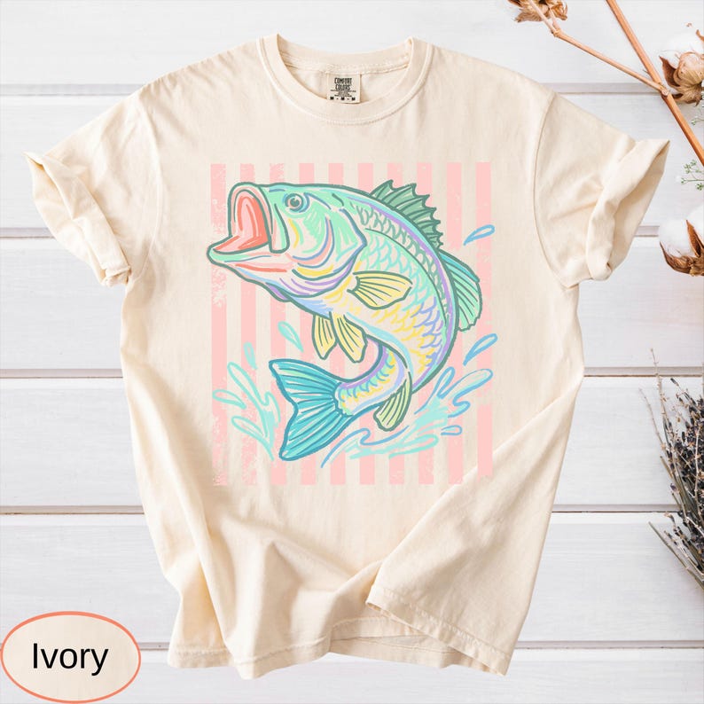Puede incluir: Camiseta de color marfil con una ilustraci&oacute;n colorida de un pez saltando con la boca abierta, salpicaduras de agua y un fondo a rayas rosas. El pez es en tonos de azul, verde, amarillo y morado. La camiseta tiene cuello redondo y mangas cortas.