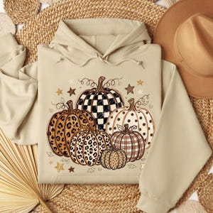 May include: A beige hooded sweatshirt featuring a graphic print of five pumpkins with various patterns including black and white checkered, leopard print, polka dots, and plaid, accented with small brown and gold stars and swirling vine illustrations.