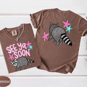 May include: Two espresso-colored t-shirts with raccoon graphics and the text "See Ya Soon." One shirt shows the raccoon with the text and stars. The other shirt shows the raccoon from the back with stars.