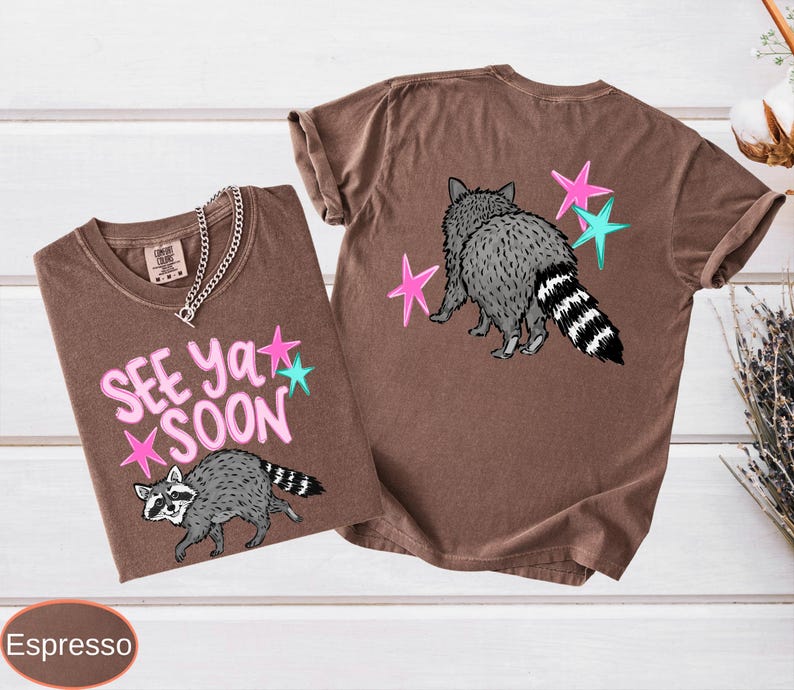 May include: Two espresso-colored t-shirts with raccoon graphics and the text "See Ya Soon." One shirt shows the raccoon with the text and stars. The other shirt shows the raccoon from the back with stars.