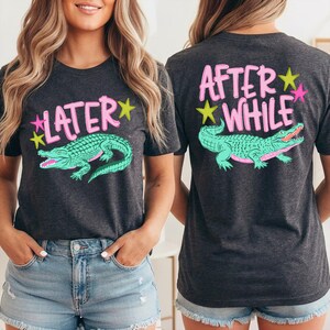 May include: Dark gray t-shirt with a front graphic of a green alligator and the word "LATER" in pink. The back of the shirt features the words "AFTER WHILE" and a matching alligator design. Yellow and pink stars accent the text.