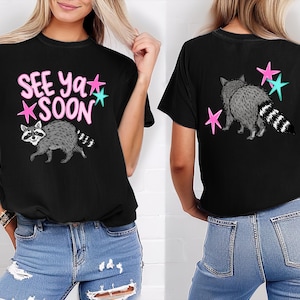 May include: Black t-shirt featuring a raccoon graphic with the text "SEE Ya SOON" in pink and teal stars. The back of the shirt shows a raccoon with pink and teal stars. The shirt is made of a soft material.