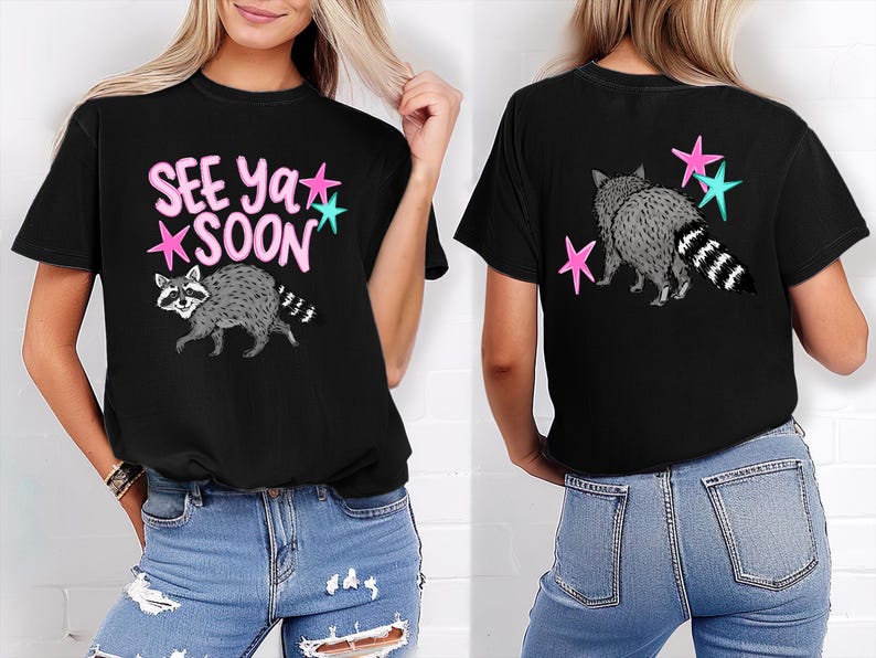 May include: Black t-shirt featuring a raccoon graphic with the text "SEE Ya SOON" in pink and teal stars. The back of the shirt shows a raccoon with pink and teal stars. The shirt is made of a soft material.