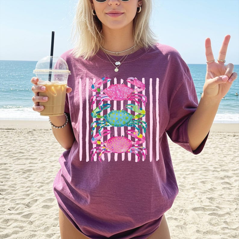May include: A maroon t-shirt featuring a graphic of three colorful crabs stacked vertically against a background of white vertical stripes. The crabs are pink, teal, and magenta. The shirt is a relaxed fit.