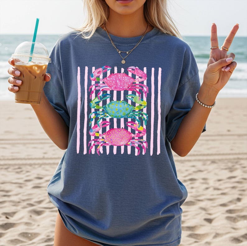 May include: A blue t-shirt featuring a design of three crabs in pink and teal, set against a backdrop of vertical pink and white stripes. The crabs have detailed patterns. The shirt is a relaxed fit.