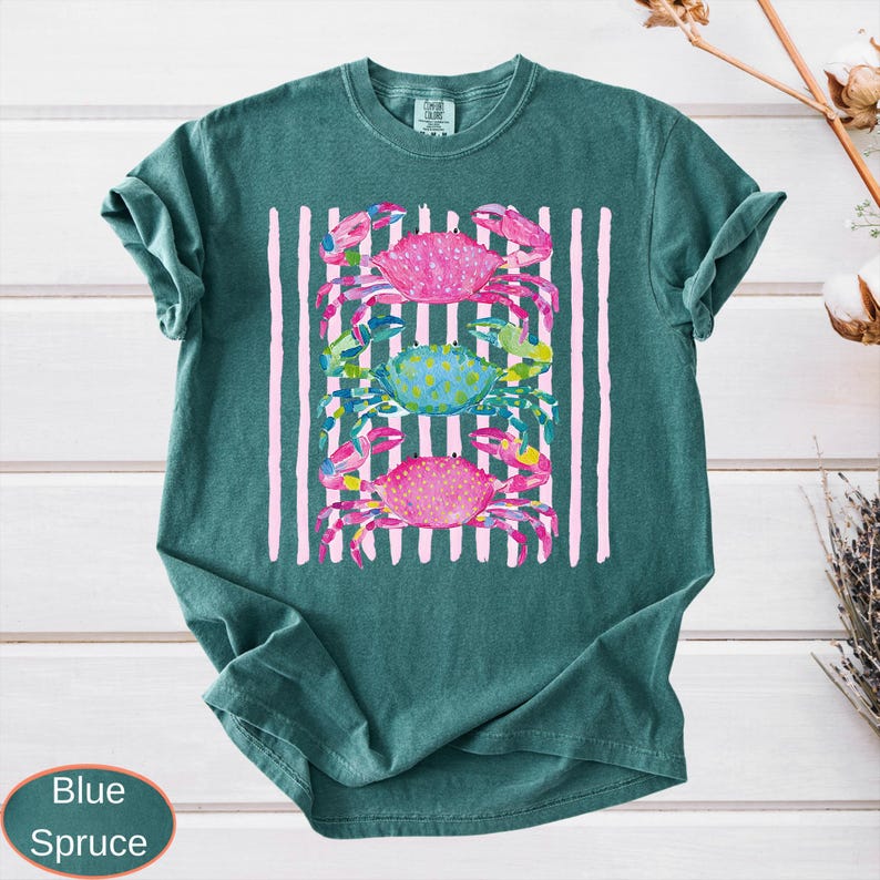 May include: A teal-colored t-shirt featuring a graphic of three crabs in pink and blue, set against a backdrop of vertical pink stripes. The crabs have detailed watercolor designs. The shirt has a relaxed fit.