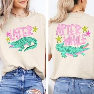 May include: Beige t-shirt with a teal alligator graphic and the word "LATER" in pink on the front. The back of the shirt features the text "AFTER WHILE" and a matching alligator design. Green and pink star accents.