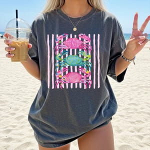 May include: Dark gray t-shirt featuring a colorful crab design. The graphic includes three crabs in pink and blue, set against a backdrop of vertical pink and white stripes. The shirt is a casual, oversized style.