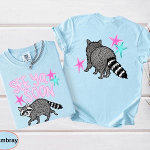 May include: Two light blue t-shirts with raccoon graphics. One shirt has the text "SEE YA SOON" in pink with pink and teal stars. The other shirt features a raccoon graphic on the back with pink and teal stars. Both shirts are made of a soft material.