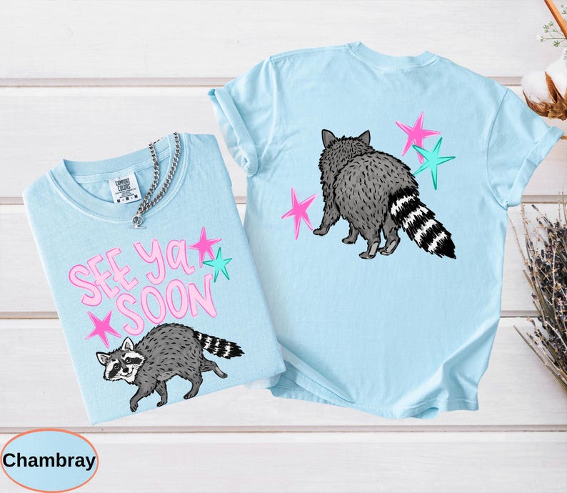 May include: Two light blue t-shirts with raccoon graphics. One shirt has the text "SEE YA SOON" in pink with pink and teal stars. The other shirt features a raccoon graphic on the back with pink and teal stars. Both shirts are made of a soft material.