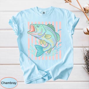 Puede incluir: Camiseta azul claro con una ilustraci&oacute;n colorida de un pez saltando con la boca abierta. El pez est&aacute; representado en tonos de verde, amarillo y azul, sobre un fondo de rayas verticales rosas. La palabra "Chambray" est&aacute; en un c&iacute;rculo en la parte inferior.